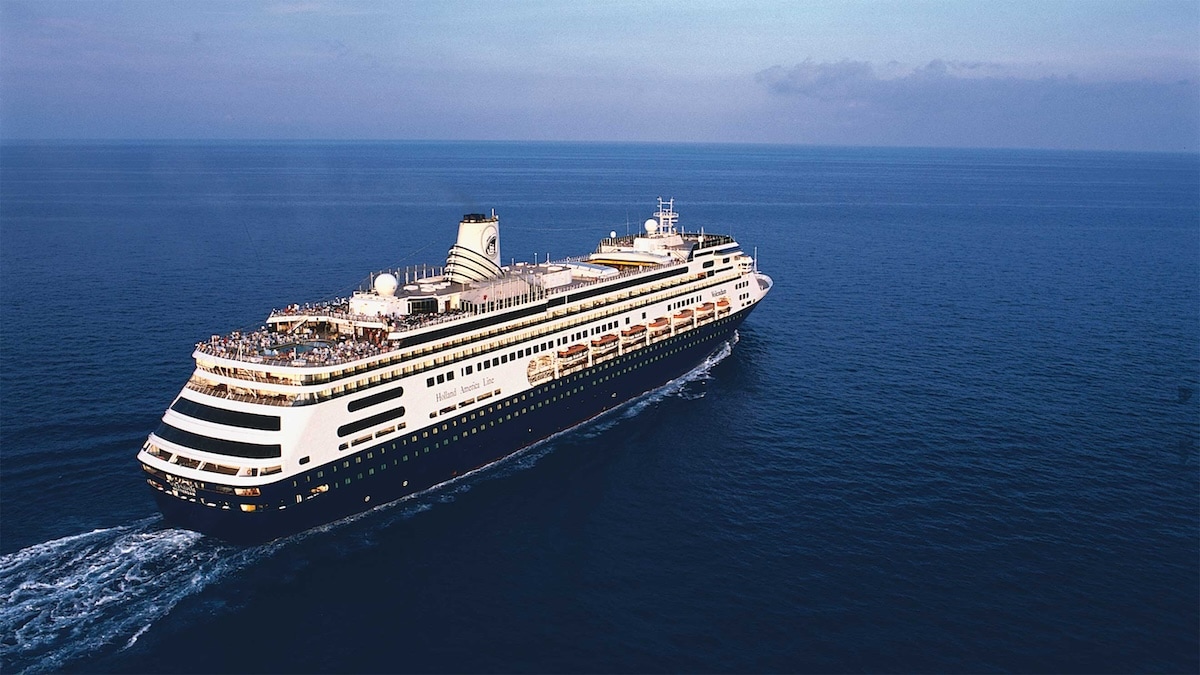 An aerial view of a large cruise ship sailing in the open ocean. The ship is white with black accents and has multiple decks. Passengers are visible on the upper decks, enjoying the sea view and outdoor activities during their 132-day World Cruise. The sky is clear, and the water is calm.