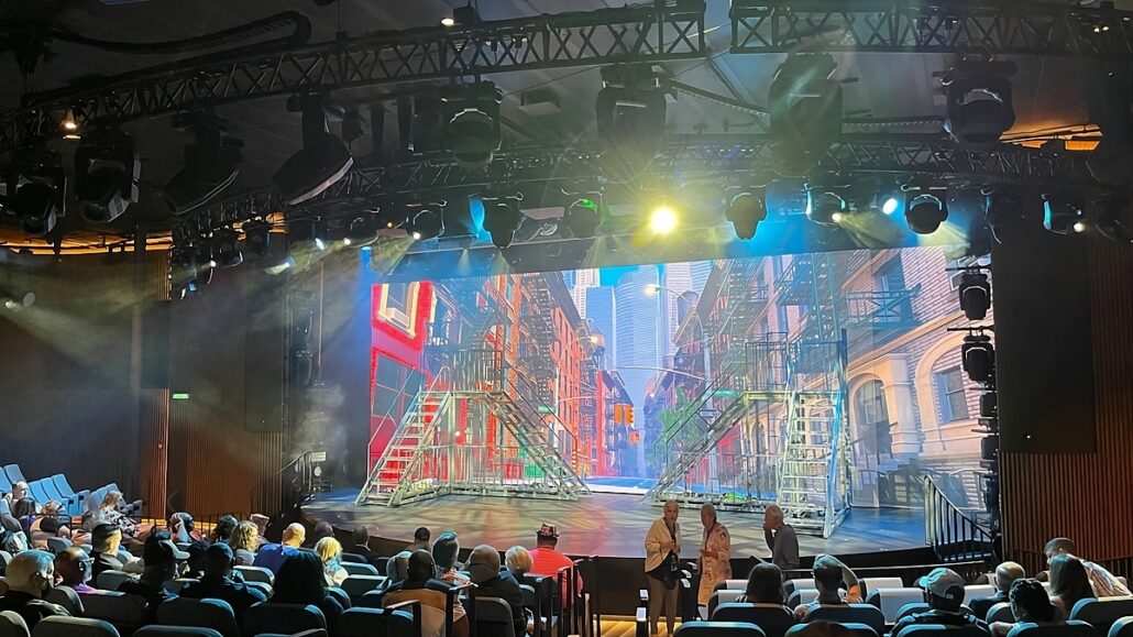 A theater stage with a vibrant city street backdrop featuring fire escapes and buildings is pictured. The audience seats are partially filled with people. Four individuals are standing near the front of the stage. Stage lights are illuminating the scene.