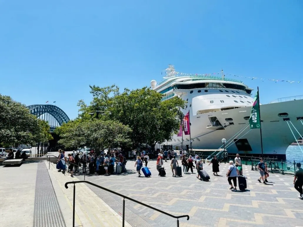 A large Royal Caribbean cruise ship docked at a port near trees, with people standing and sitting on benches nearby. Some people are seen with suitcases, likely affected by technical glitches that cancels sailings. There is a bridge in the background under a clear blue sky.