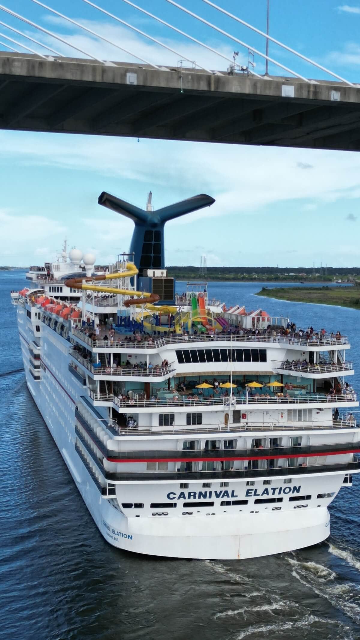 A large white cruise ship named "Carnival Elation" is sailing under a bridge, departing from the Florida Cruise Port. The ship, crowded with people, boasts various amenities including seating areas, pools, and water slides. The sky is clear and the surrounding water is calm.