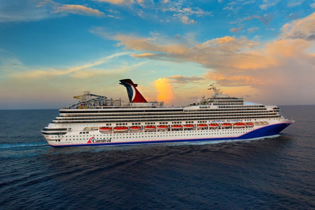 A large Carnival Cruise ship sails on the open sea under a colorful sunset sky. The vessel, featuring multiple decks and a distinctive red and blue funnel, moves smoothly across the water.