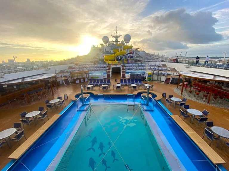 A view of the Carnival Sunrise deck featuring a swimming pool with dolphin designs, lounge chairs, dining tables, and sun loungers. In the background are water slides, and the sun is setting behind a partly cloudy sky. Perfect for photos before its 2024 dry dock.