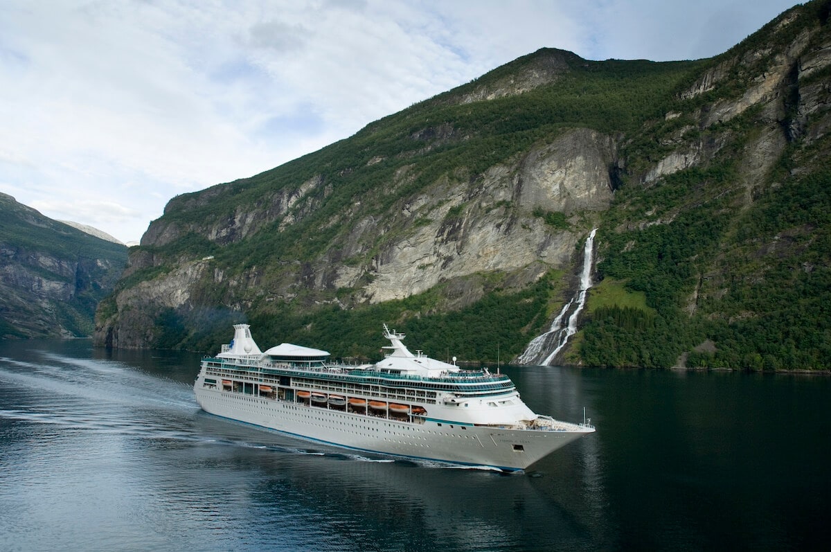 A Royal Caribbean cruise ship sailing near a waterfall along a fjord with steep mountainsides.