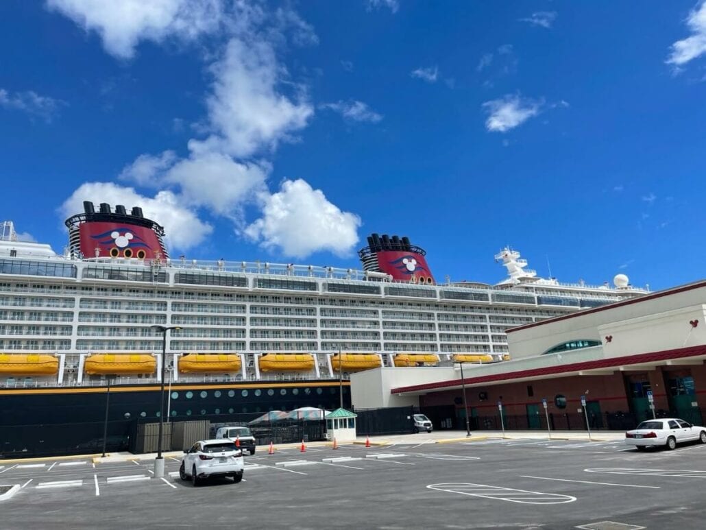 A disney cruise ship docked at a port with a clear blue sky above, and a nearly empty parking lot in the foreground.