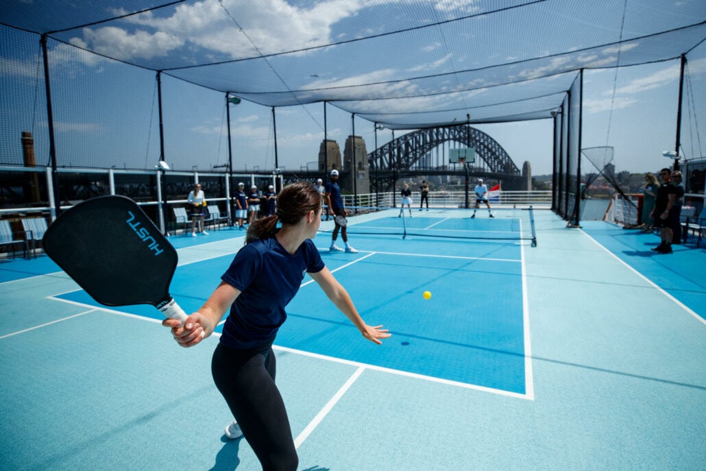 A person with brown hair in a ponytail, wearing a navy blue shirt and black leggings, prepares to hit a yellow ball with a paddle on one of the well-lit pickleball courts on a cruise ship. The court is surrounded by netting and offers a view of the ocean and distant bridge in the background.