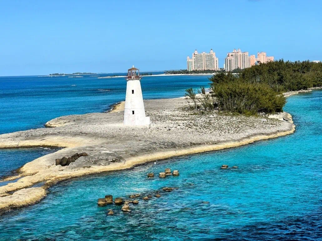A white lighthouse stands on a small rocky island in Nassau, Bahamas surrounded by turquoise water. In the background, Nassau reveals a cluster of tall, pink buildings amid greenery under a clear blue sky.