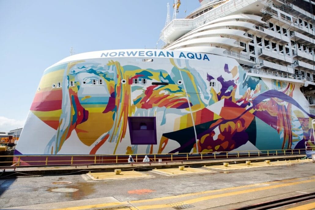 Colorful abstract mural on the bow of the norwegian aqua cruise ship, featuring whale designs, docked in a port with visible workers.