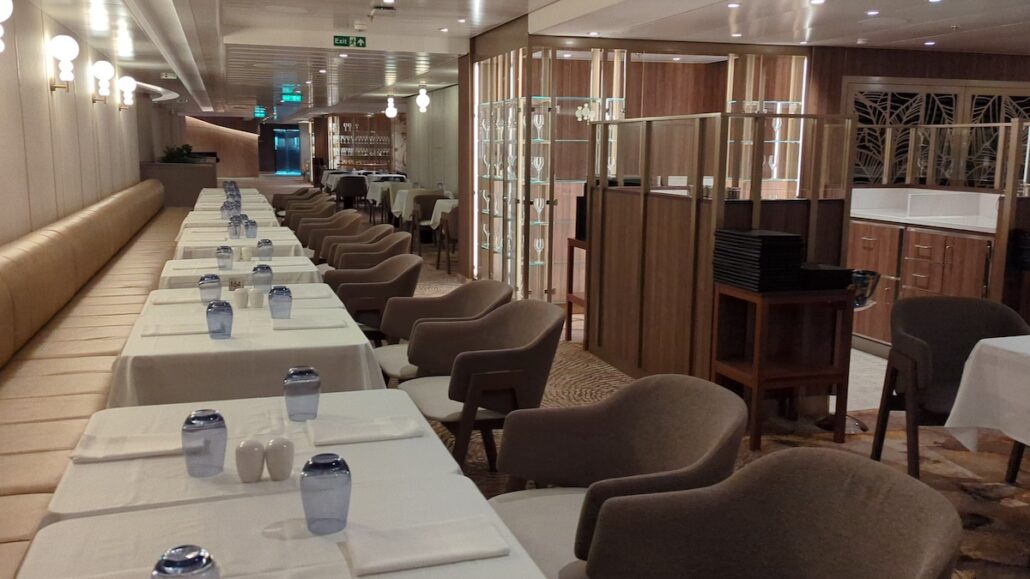 A spacious, modern dining area with rows of tables and chairs set neatly. Each table has a white tablecloth, blue water glasses, and salt and pepper shakers. The warm lighting and wooden paneling give the space an elegant atmosphere, perfect for enjoying Princess Cruises' flexible dining options.