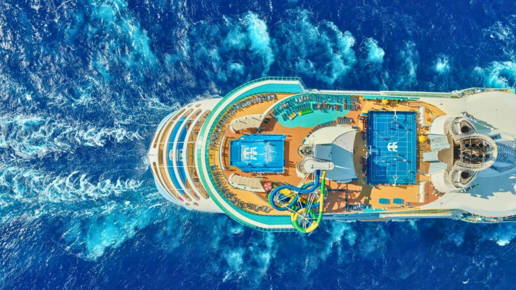Top view of a cruise ship's deck featuring pools, water slides, and sports courts sailing on blue ocean waters as a cruise passenger jumps overboard in front of family.