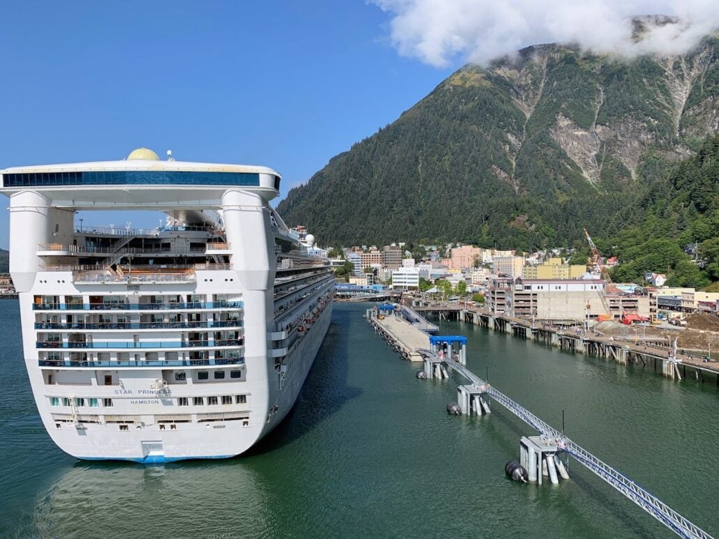 A large cruise ship, managed by prominent cruise lines, docked at a port with a small town and mountainous backdrop under a clear blue sky.