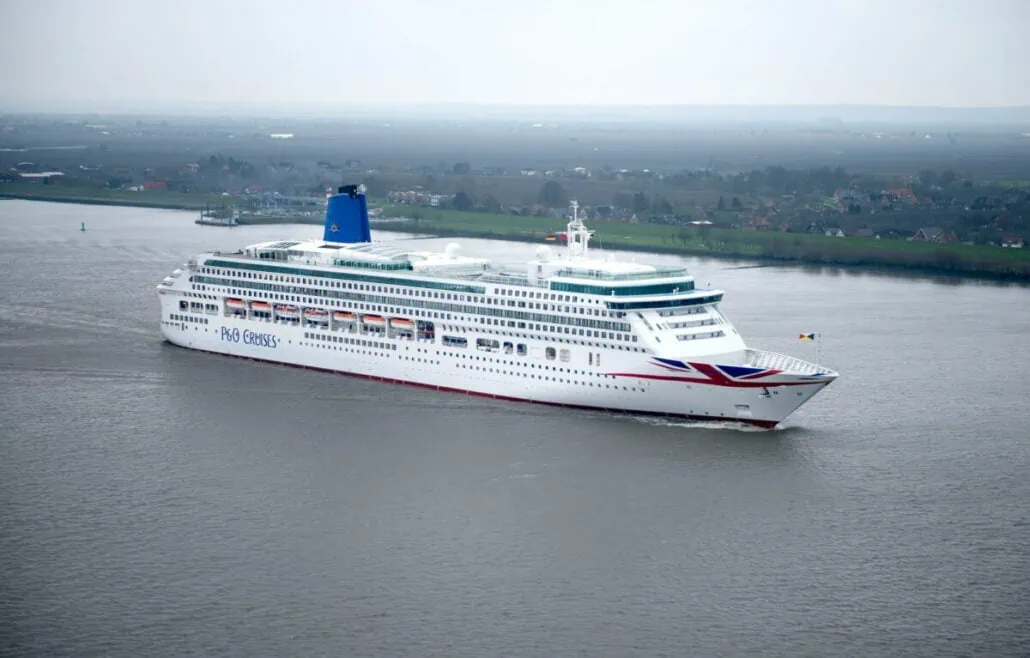 A large white P&O cruise ship from the UK sails on a wide river with a vast landscape and scattered buildings in the background under a cloudy sky.