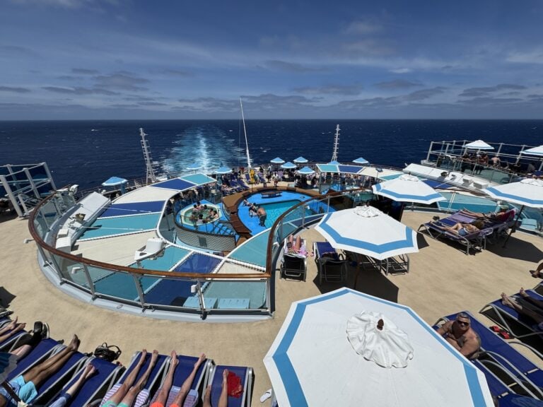 View from the deck of a cruise ship showing people relaxing by a circular pool with lounge chairs and parasols. The ocean is visible in the background, along with the ship's wake trailing behind. The sky is clear with a few scattered clouds.
