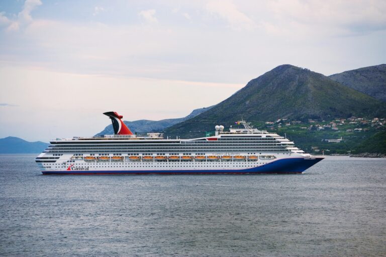 A large Carnival cruise ship sails on calm waters, evoking the lively spirit of New Orleans. Green hills and scattered buildings form a picturesque backdrop under an overcast sky.