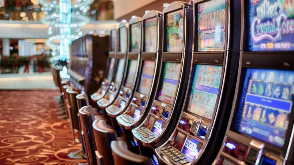 A row of colorful slot machines lines the casino on the cruise, set against a red and gold patterned carpet with chandeliers gleaming above. The machines, aglow with bright lights, showcase diverse game themes as passengers try their luck amidst dreams of winning big.