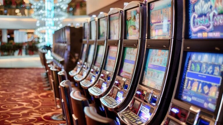 A row of colorful slot machines lines the casino on the cruise, set against a red and gold patterned carpet with chandeliers gleaming above. The machines, aglow with bright lights, showcase diverse game themes as passengers try their luck amidst dreams of winning big.