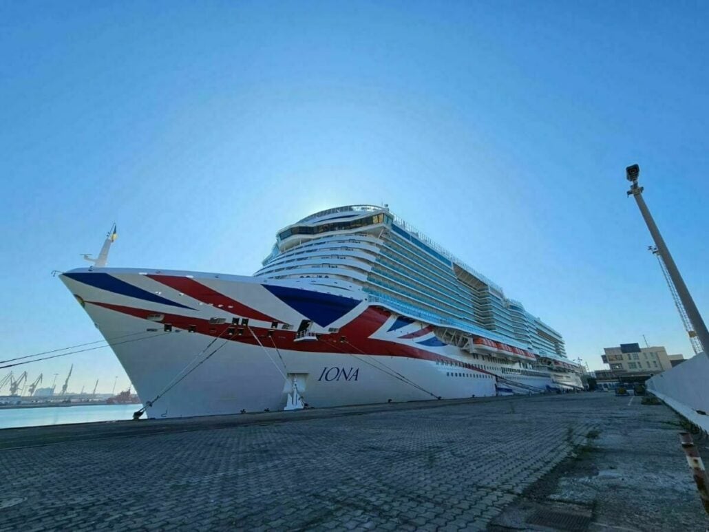 A large white P&O cruise ship named "Iona" docked at a UK port, featuring a bold Union Jack design on its bow. Clear blue sky overhead highlights its impressive size.