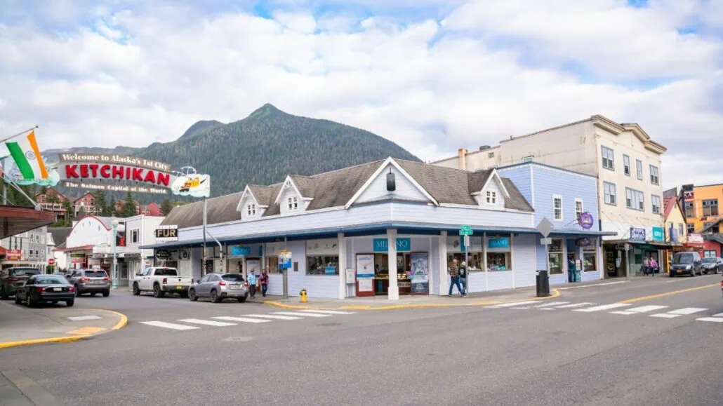 A street view in Ketchikan, Alaska, featuring several shops and buildings under a cloudy sky with mountains in the background. A welcome sign reads, "Welcome to Alaska's First City, Ketchikan," where a 91-year-old tourist had a minor scooter accident last year.
