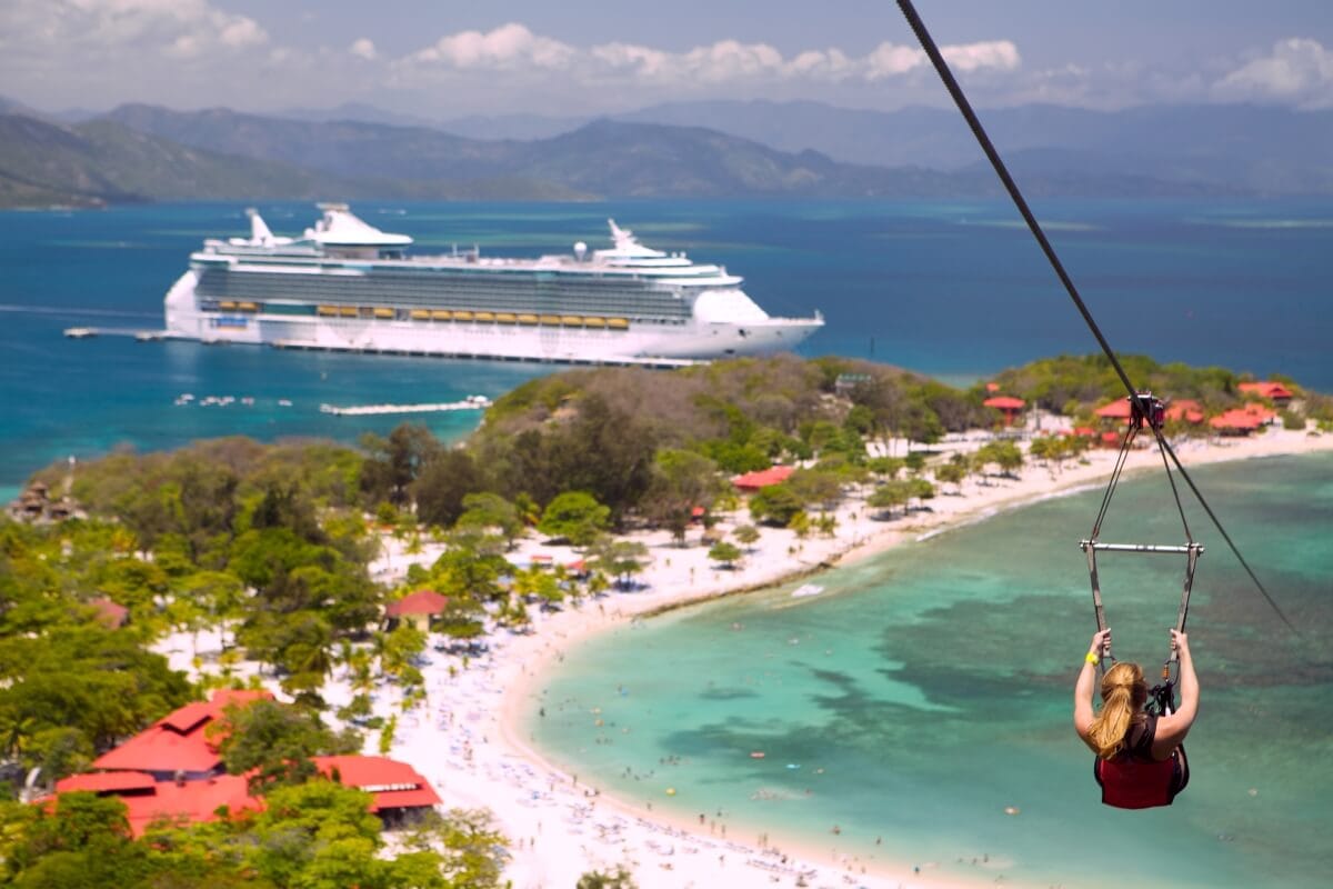 A person rides a zipline over a beach with a large Royal Caribbean cruise ship docked near Labadee Island in the background.