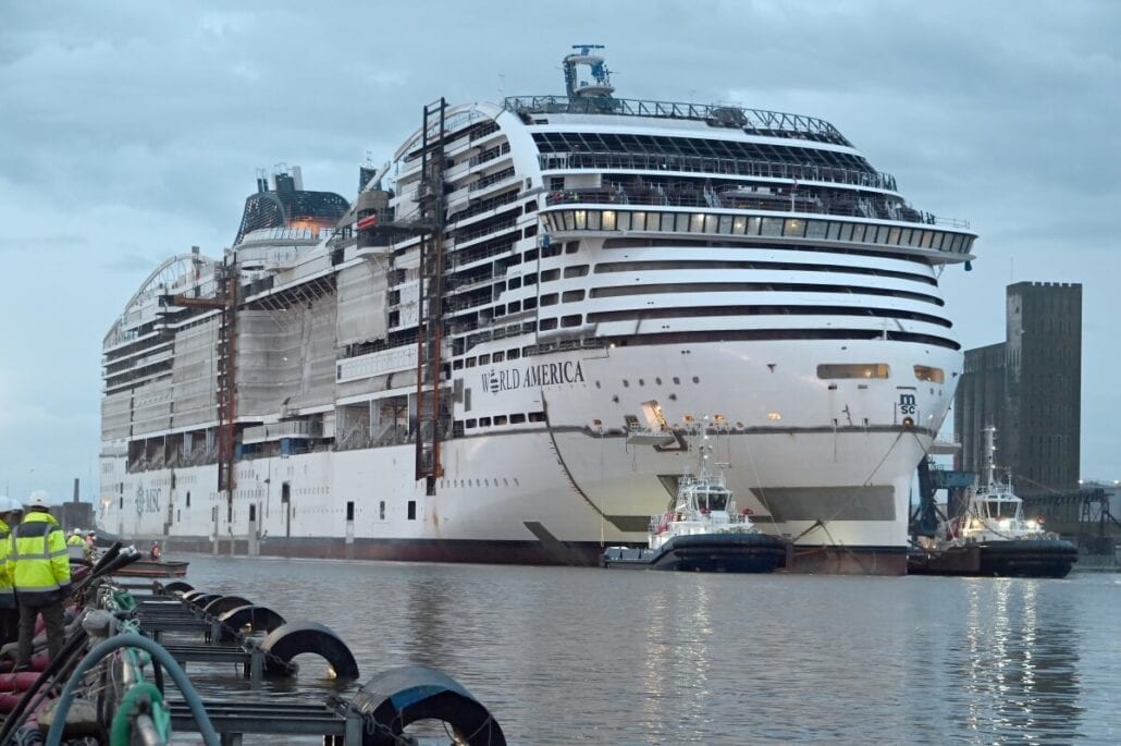 A large cruise ship named "World America," commissioned by MSC Cruises, is being constructed and fitted at Meyer Turku shipyard, surrounded by cranes and workers on a cloudy day.