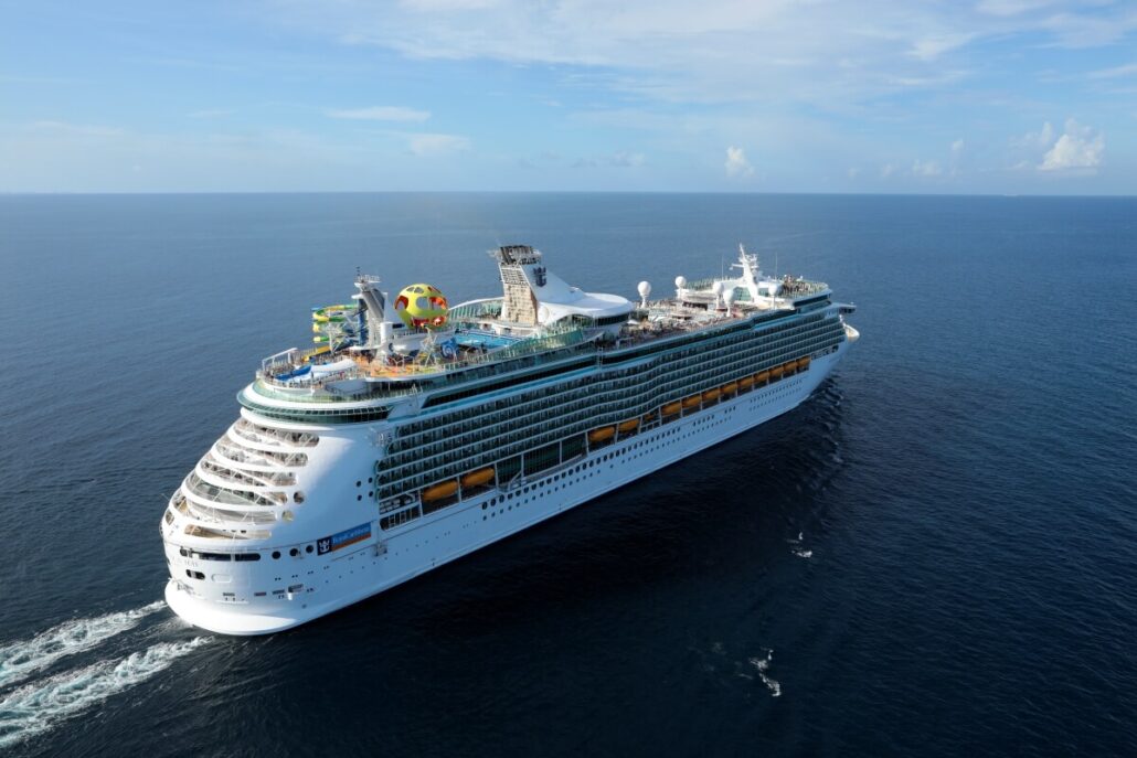 The Mariner of the Seas, a large cruise ship, sails on the ocean under a clear blue sky. This majestic vessel features multiple decks, a water slide, and various recreational areas as it glides through the Western Caribbean's open sea with no land visible in the background.
