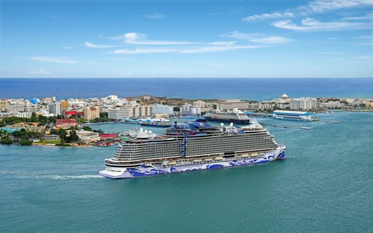 A large Norwegian ship departing from a coastal city in Spain with colorful buildings and clear blue skies.
