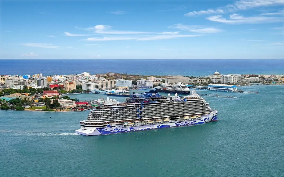 A large Norwegian ship departing from a coastal city in Spain with colorful buildings and clear blue skies.