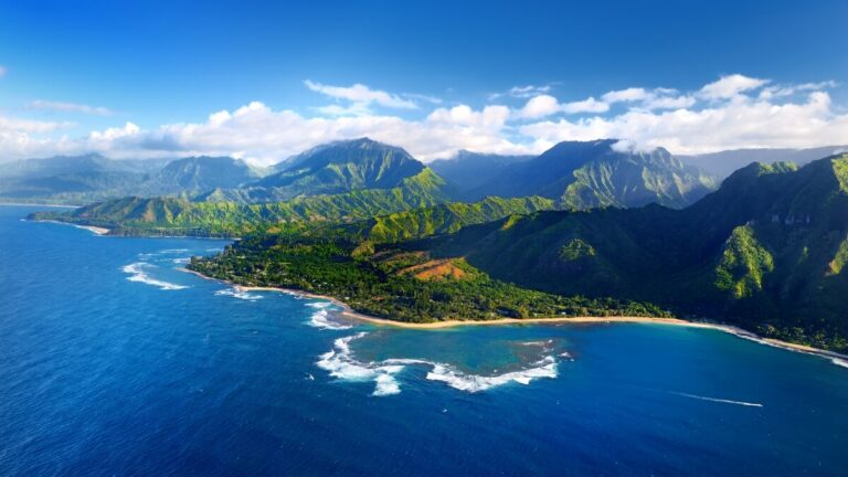 Aerial view of the lush, mountainous Nā Pali Coast with clear blue waters and waves crashing on the shore.