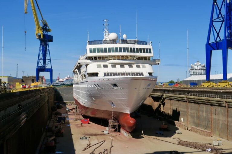 A large cruise ship sits in a dry dock for maintenance under clear blue skies, with cranes and various equipment visible around it. Due to water tank issues, the world cruise has been delayed.