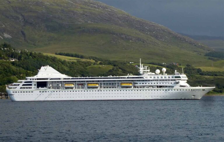 A large white cruise ship from Villa Vie Cruises is anchored near the shore, with green hills and a mountain in the background, ready to welcome passengers eager to explore or perhaps even retire at sea.