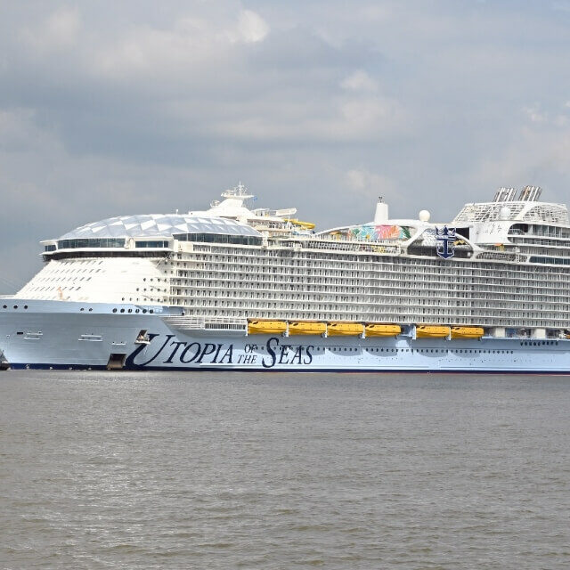 Large cruise ship, "Utopia of the Seas," embarks on water with two escorting tugboats under a cloudy sky.