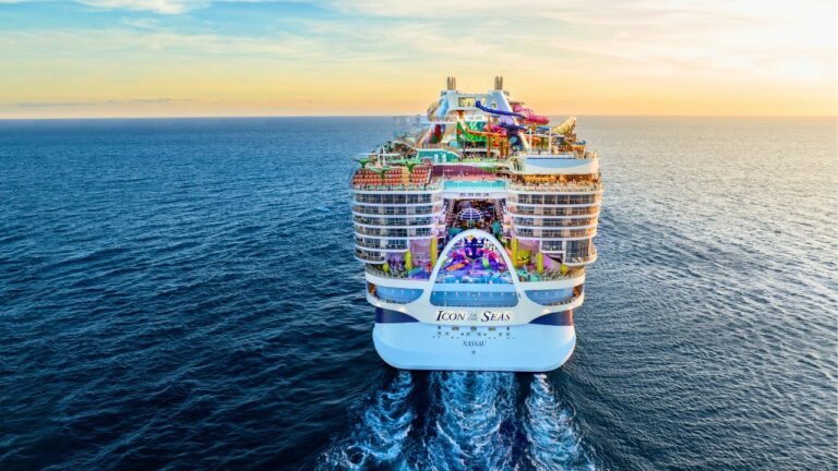 The world's largest cruise ship, "Icon of the Seas," sails on the ocean at sunset, with colorful lights and amenities visible on its decks.