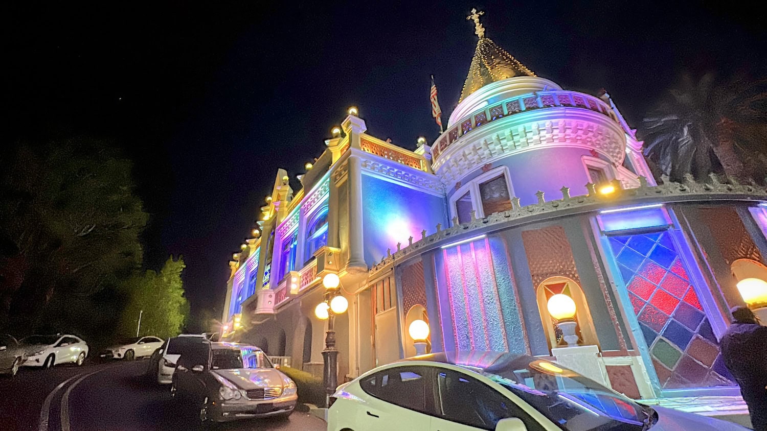 A brightly lit building with domes and colorful lights at night hints at hidden secrets. Several cars are parked along the curved street in front, while trees are visible on the left side.