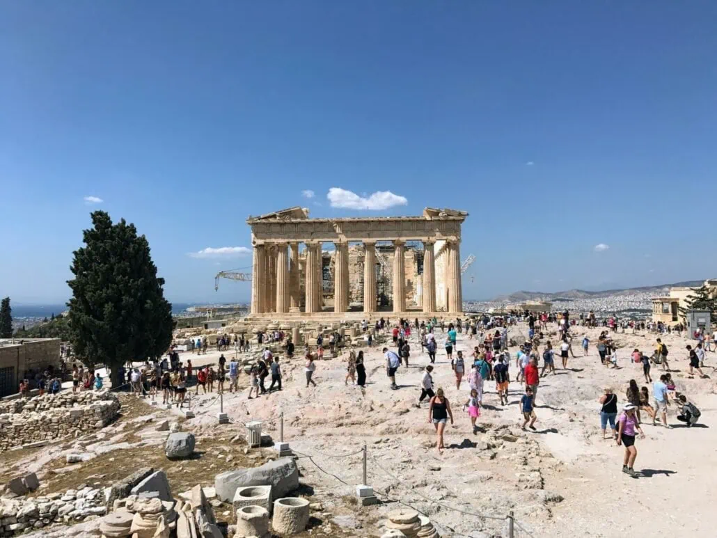 Tourists explore the area in front of the Parthenon in Athens, Greece, with clear skies overhead and scattered ancient ruins in the foreground, as cruisers are warned about the extreme heatwave impacting the region.