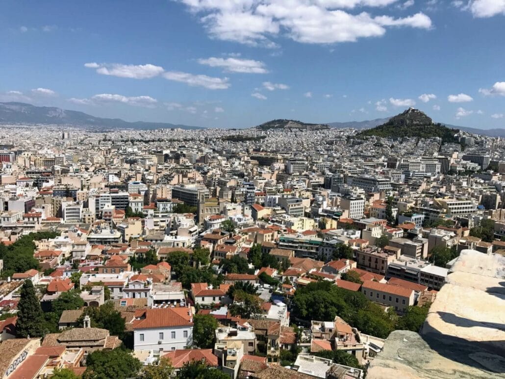 Aerial view of a sprawling cityscape in Greece, with densely packed buildings, red-tiled roofs, and a distant hill under a partly cloudy sky. Amid the extreme heatwave, cruisers dot the nearby coastline.