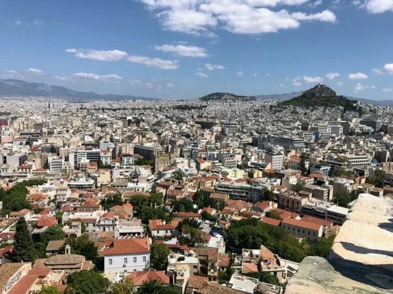 Aerial view of a sprawling cityscape in Greece, with densely packed buildings, red-tiled roofs, and a distant hill under a partly cloudy sky. Amid the extreme heatwave, cruisers dot the nearby coastline.