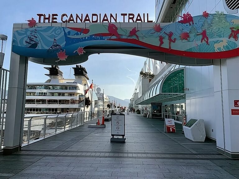 Entrance to The Canadian Trail with colorful arch, illustrations, and signage. A large cruise ship docked at the pier for embarkation in Vancouver. A few cones and signs present. Clear sky and distant mountains in the background.