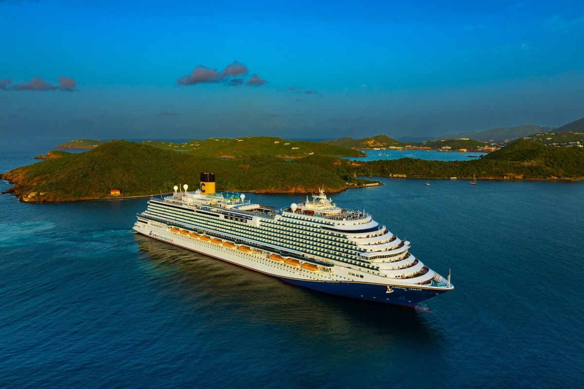 A large cruise ship sails through calm blue waters near green, hilly islands under a clear sky.