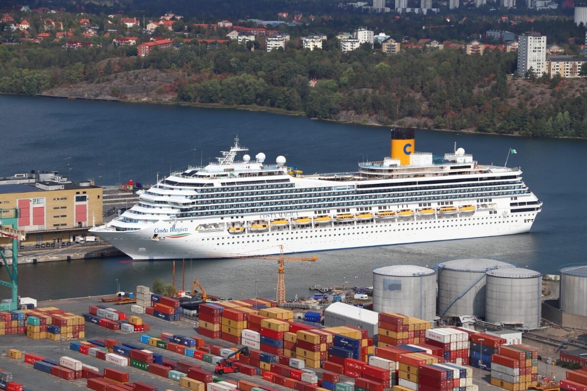 A large white cruise ship docked at an industrial port, surrounded by numerous shipping containers and storage tanks, with a wooded area and cityscape in the background.