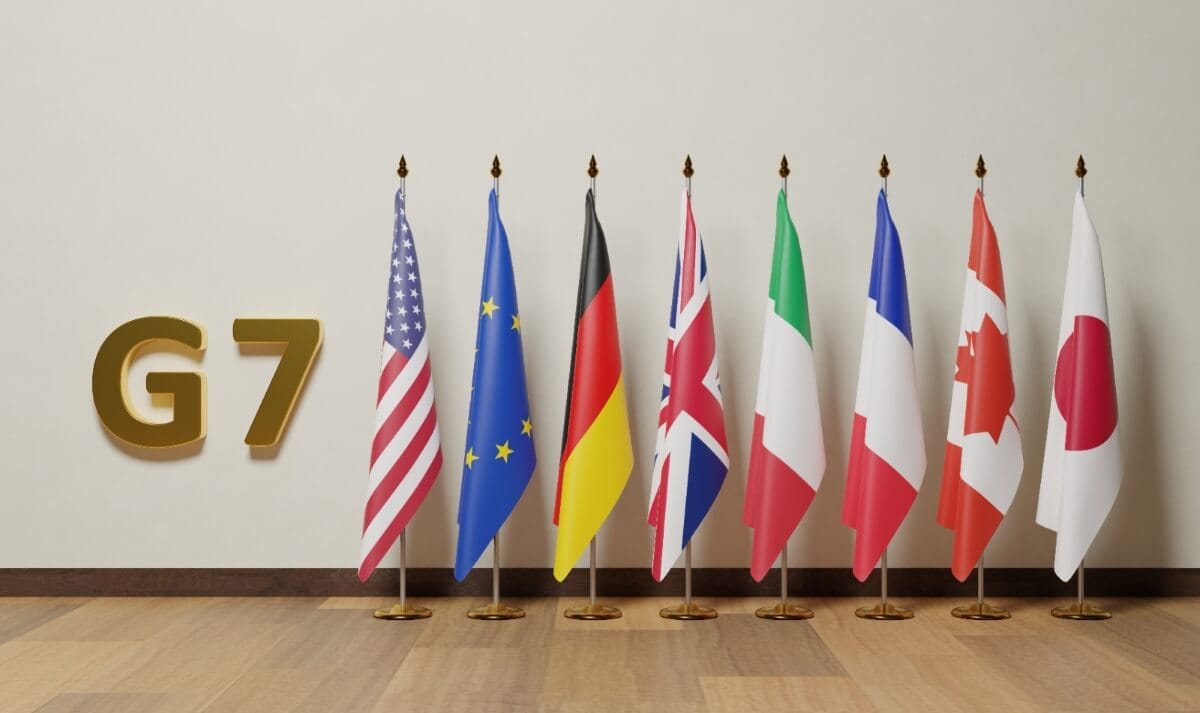 Flags of the G7 member countries (United States, European Union, Germany, United Kingdom, Italy, France, Canada, and Japan) are displayed on flagpoles in a row with a 'G7' sign on the left.