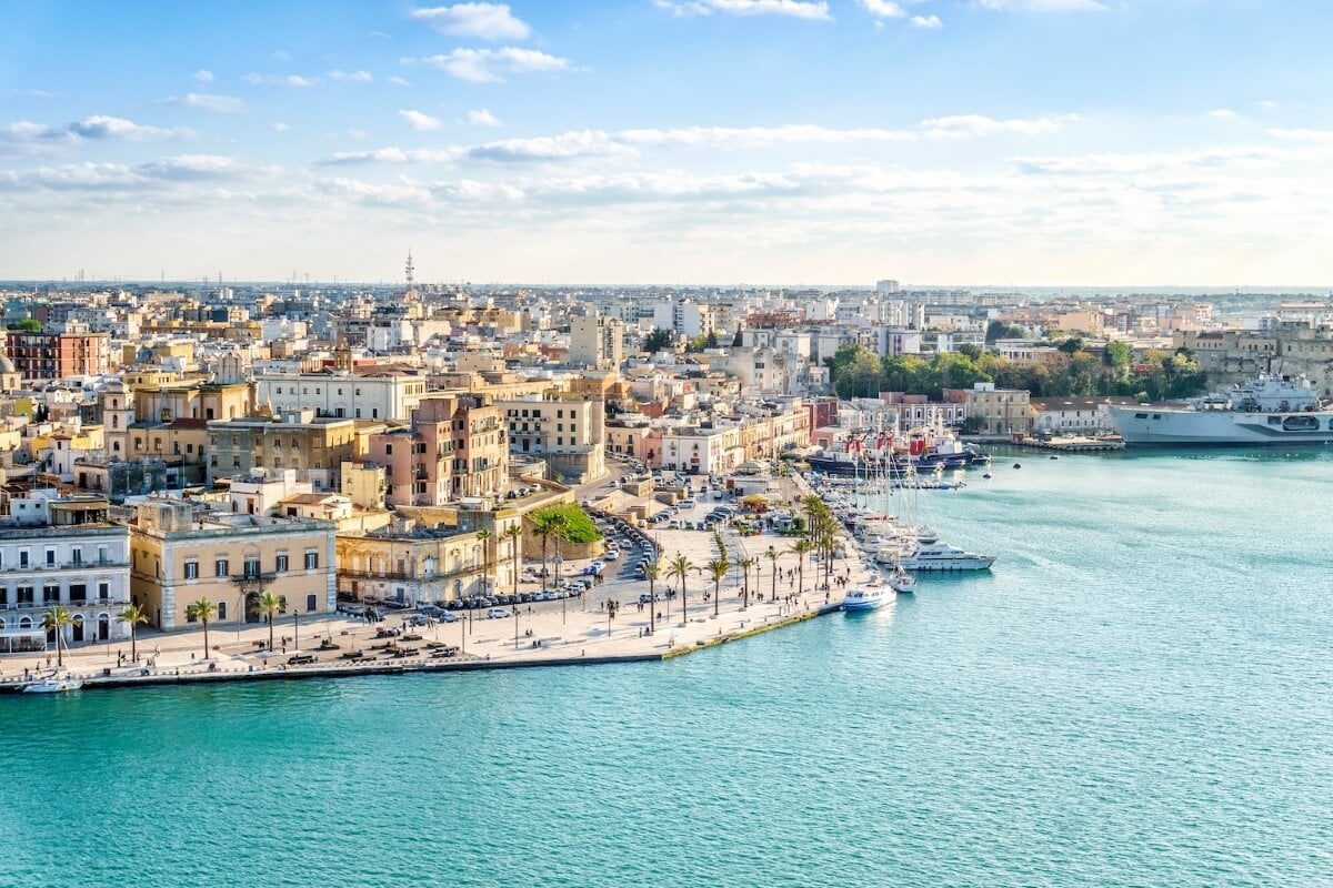 A coastal cityscape featuring historic buildings, a marina with docked boats, and a clear blue sky over turquoise waters.