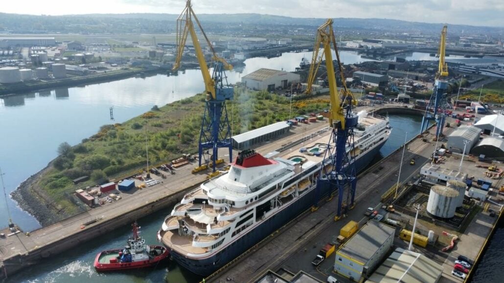 Aerial view of a large ship docked in a shipyard with two yellow cranes nearby, a tugboat alongside, and industrial buildings in the background, preparing for its 300-day cruise.