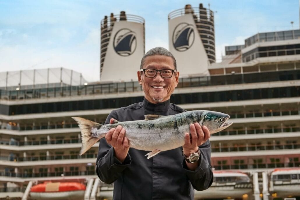 A smiling man in a black shirt holds a large fish in front of a cruise ship.