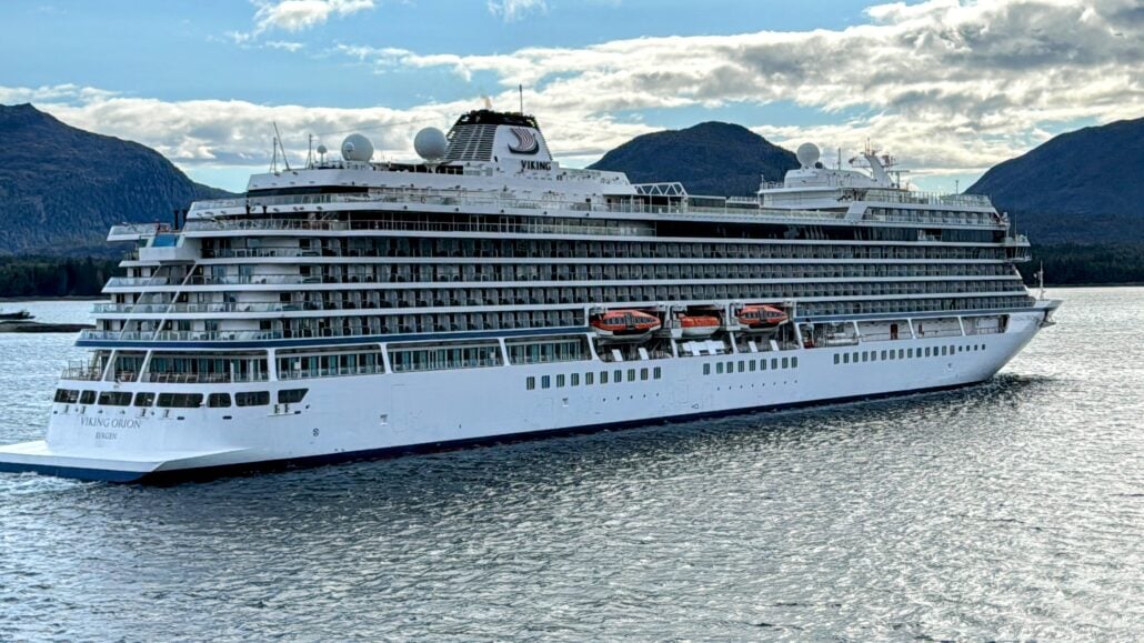 A large white ocean cruise ship, reminiscent of the majestic Viking fleet, sails on calm water with mountainous terrain in the background under a partly cloudy sky.