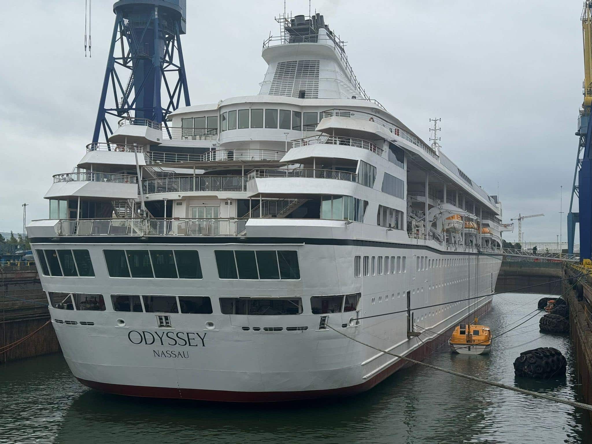 A large world cruise ship named "Odyssey Nassau" docked in a shipyard in Belfast. A small yellow boat floats nearby in the water, possibly a sign of technical delays.