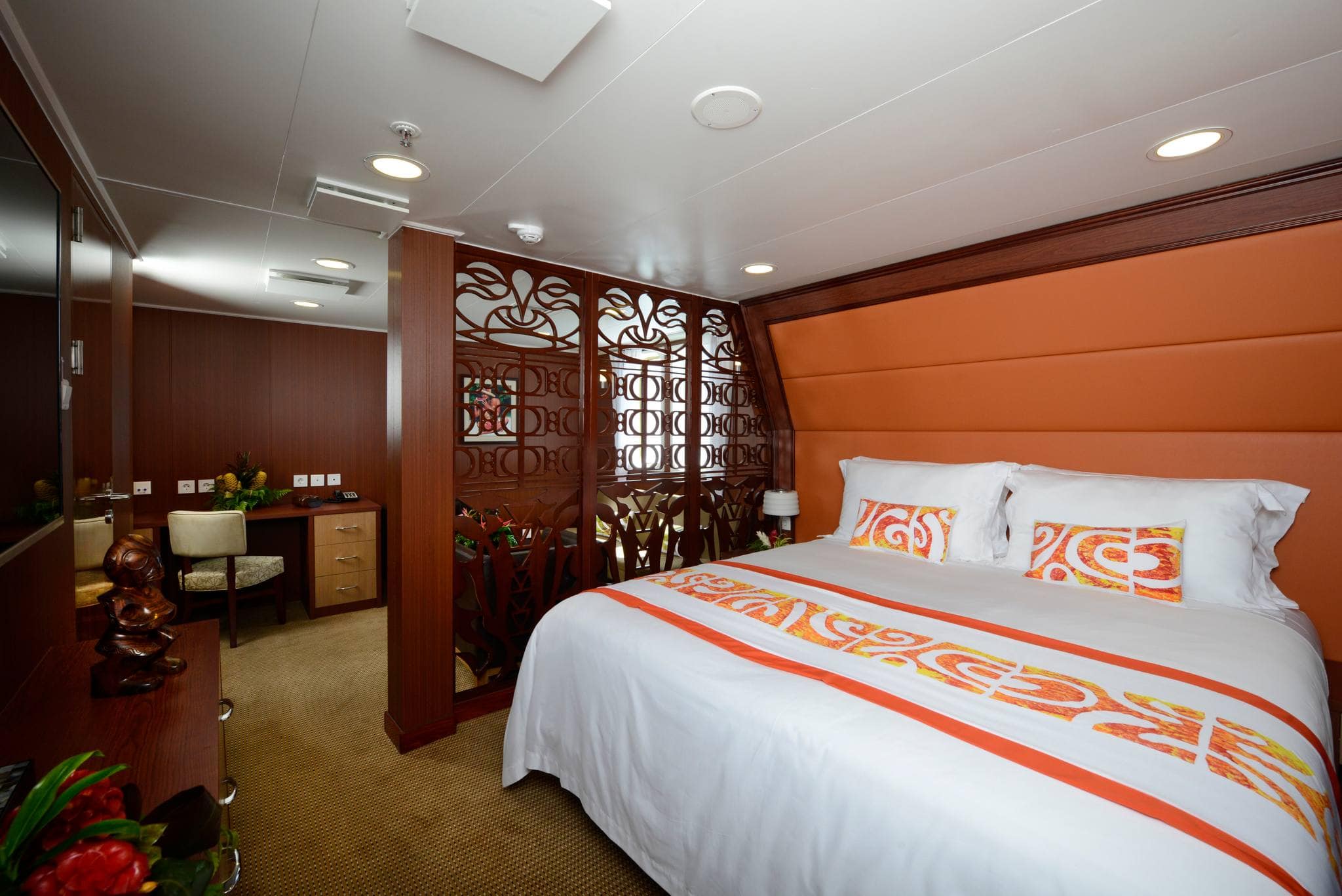 A modern cruise ship cabin with a bed, decorative screen, and desk. The bed has white linens and orange accents. The cabin has dark wood paneling and an orange padded wall. Various decor items are visible, making it feel like a luxurious room in the floating Olympic village.