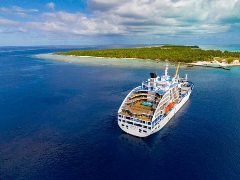 A large cruise ship, resembling a floating Olympic village, sails in calm blue waters near a tropical island with lush greenery and white sandy shores under a partly cloudy sky.