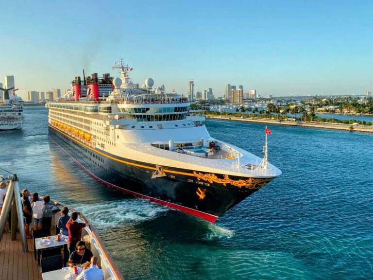 All aboard a large cruise ship sailing through a scenic waterway with a city skyline in the background. Passengers on board another Disney vessel watch and take photos, capturing the magical moment.