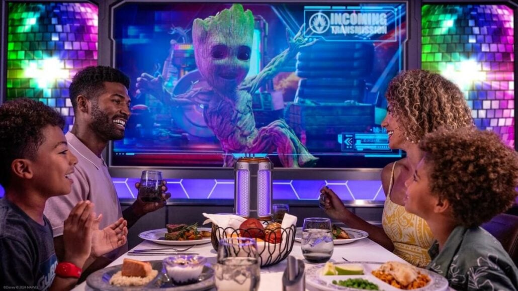 A family of four smiles at a table in a futuristic setting, enjoying good eats while watching a virtual character, Groot, on a screen behind them labeled "Incoming Transmission." This delightful scene is part of the Disney Treasure's featured dining venues.