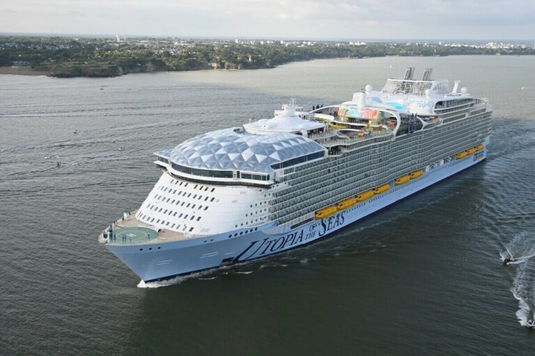 A large cruise ship named "Utopia of the Seas" sails on the ocean near a coastline, possibly en route to Port Canaveral. The ship features multiple decks, numerous amenities, and a distinctive dome structure on the top front.