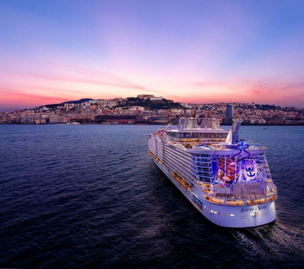A Royal Caribbean cruise ship sails on a calm sea with a coastal city and a hillside illuminated by sunset in the background, showcasing a $100M Glow-Up.